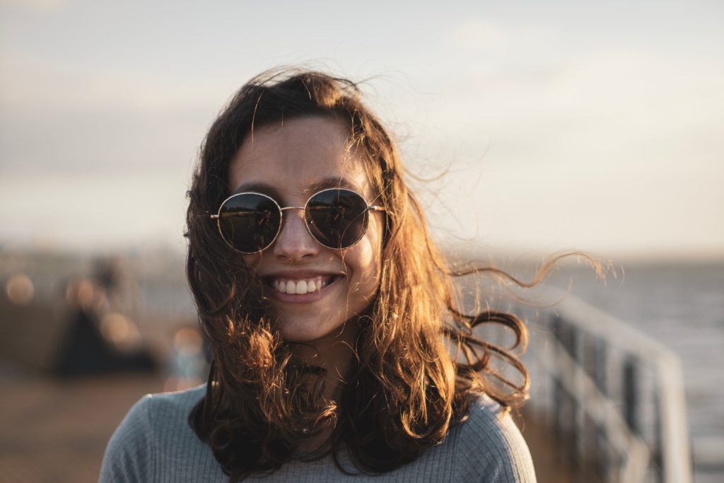 A woman with sunglasses smiling at the beach on a windy day.